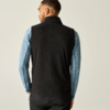Persoon van achteren, draagt zwart mouwloos fleecegilet over lichtblauw shirt met lange mouwen, voor een lichte achtergrond