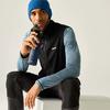Man zit op een wit blok, houdt een donkerblauwe drinkfles dicht bij zijn mond, draagt een blauwe gebreide muts, zwart vest over een blauw gestreept shirt met lange mouwen en zwarte broek