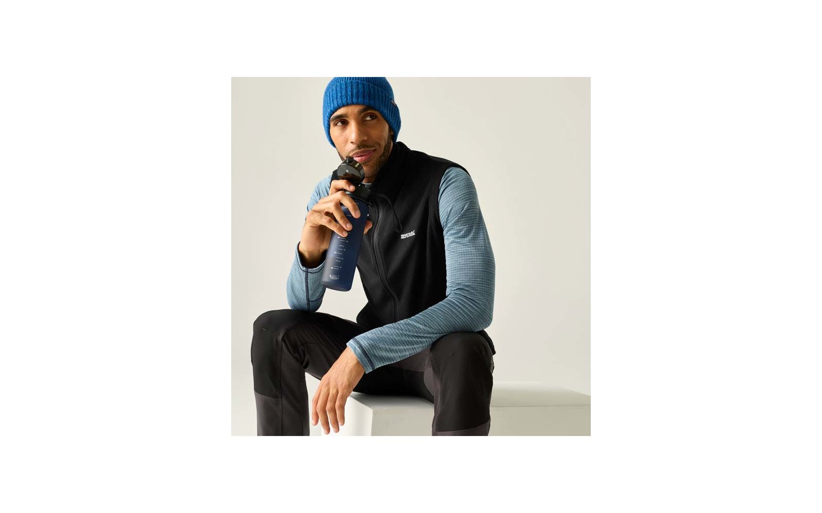 Man zit op een wit blok, houdt een donkerblauwe drinkfles dicht bij zijn mond, draagt een blauwe gebreide muts, zwart vest over een blauw gestreept shirt met lange mouwen en zwarte broek