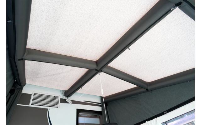 Decke mit hellen Stoffpaneelen, dunklen Querträgern, einer hängenden Schnur, einem rechteckigen Lüftungsgitter an der Wand und einem Fenster mit Rahmen