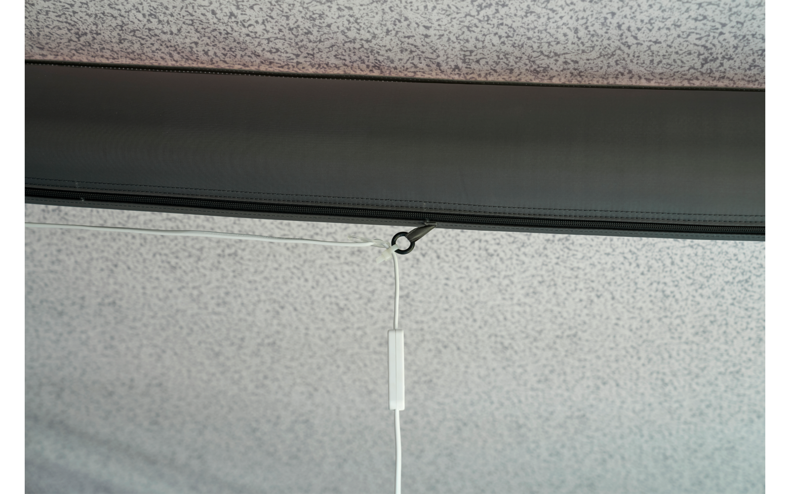 Grau gesprenkelte Decke mit einer dunklen horizontalen Leiste; an einer kleinen schwarzen Öse hängt eine weiße Schnur mit einem rechteckigen weißen Kunststoffgriff