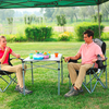 Deux personnes assises sur des chaises de camping en plein air, avec une petite table pliante et des tasses colorées entre elles