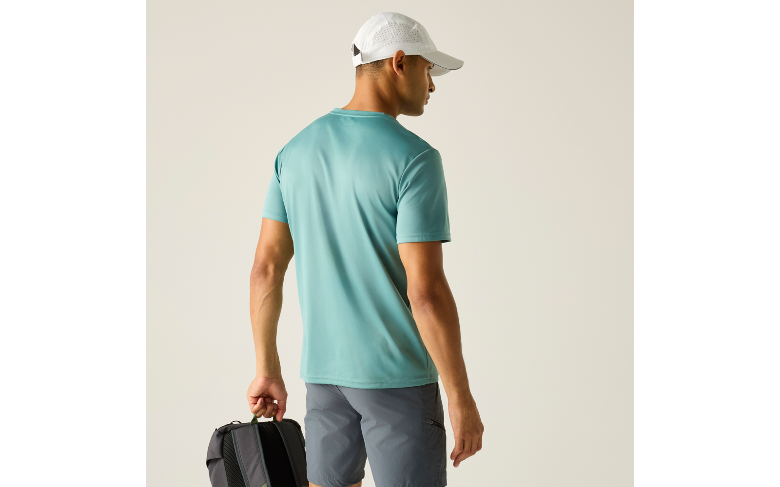 Person von hinten trägt türkisfarbenes Kurzarm-T-Shirt, graue Shorts und weiße Kappe, hält einen schwarzen Rucksack in der linken Hand