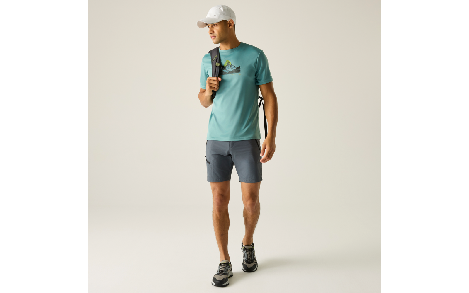 Mann mit weißer Cap, türkisem T‑Shirt mit Bergmotiv, grauen Shorts, Wanderschuhen und Rucksack über einer Schulter, gehend und nach rechts blickend