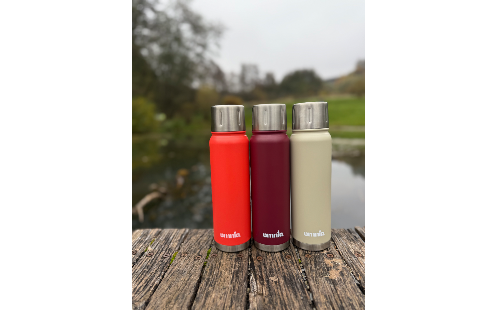 Drie flessen met roestvrijstalen doppen (rood, donkerrood, beige) naast elkaar op verweerd hout; op de achtergrond onscherpe vijver en bomen; witte logo-opdruk op de flessen