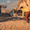 Two children lift folding camping tables on sandy ground; tent, SUV and large rock formation in the background