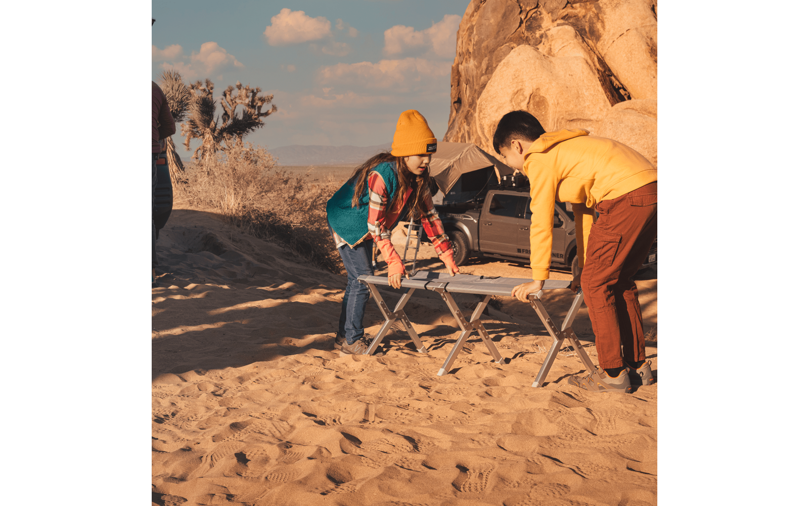 Two children lift folding camping tables on sandy ground; tent, SUV and large rock formation in the background