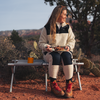 Person with long hair sits on a camping bench in a red rocky landscape, holding a plate of food and a fork, an orange cup on the bench, wearing a light fleece jacket, dark pants and brown hiking boots, looking to the right