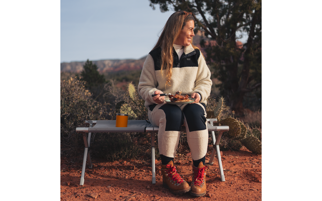 Person with long hair sits on a camping bench in a red rocky landscape, holding a plate of food and a fork, an orange cup on the bench, wearing a light fleece jacket, dark pants and brown hiking boots, looking to the right