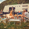 Five adults sitting and standing at a camping table in front of a motorhome on a grassy field, with plates and food on the table