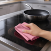 Hand mit rosa Reinigungstuch wischt schwarzes Glaskeramik-Kochfeld; Pfanne auf dem Kochfeld, Arbeitsfläche und Küchenschränke im Hintergrund