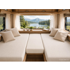 Interieur van een camper met twee beige gestoffeerde ligplaatsen en kussens, houten bovenkastjes en een groot panoramaraam met uitzicht op een meer, naaldbomen en bergen