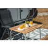 Table de camping en bois près d'une caravane avec deux verres de jus d'orange, un bol de raisins, un petit vase blanc avec une plante, deux chaises de camping pliantes et un tapis d'extérieur à motif