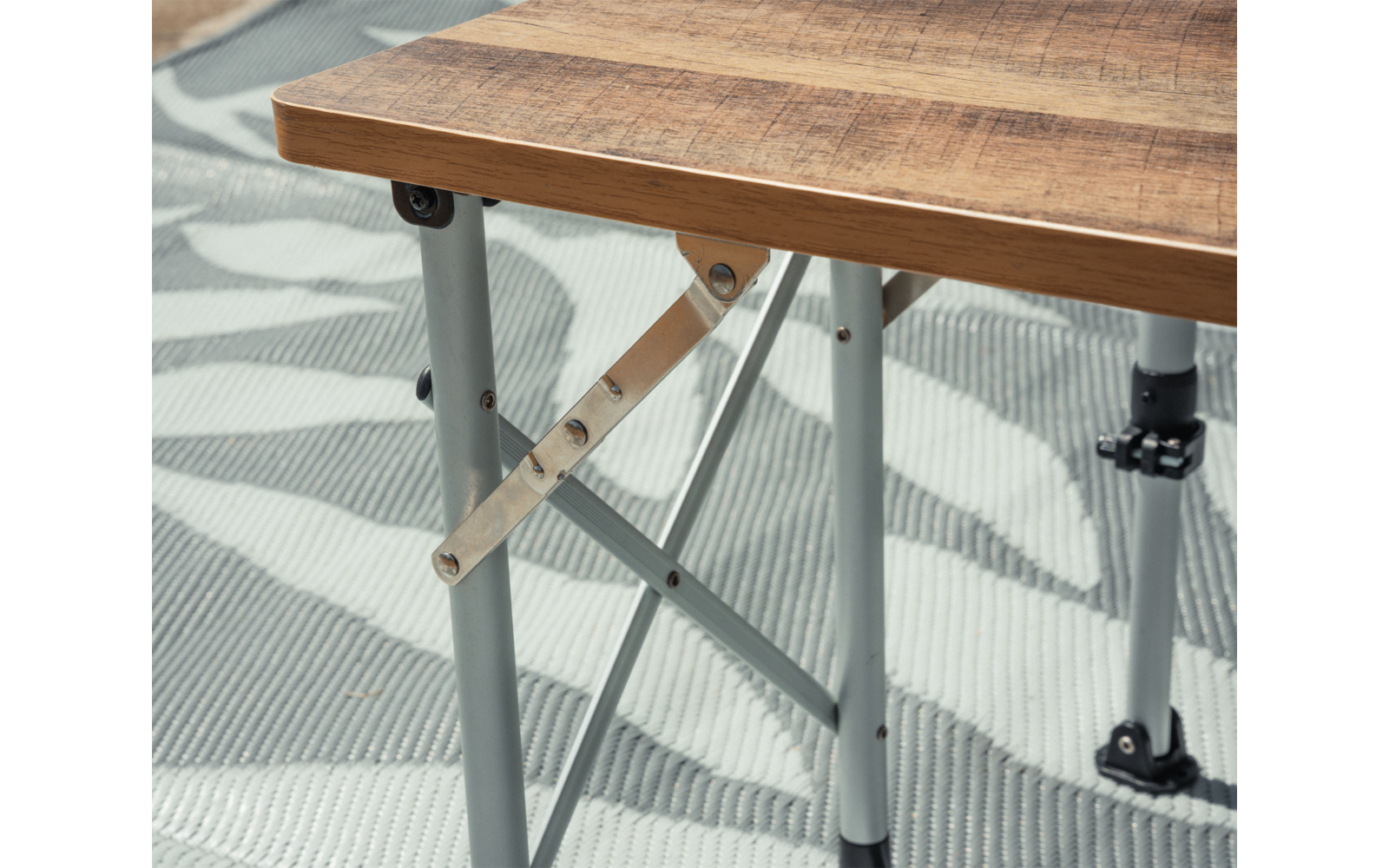 Plateau de table en bois avec pieds métalliques gris et entretoises diagonales sur un tapis gris clair à motifs de feuilles blanches