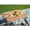 Deux tables pliantes en bois sur un tapis gris à motifs posé sur l'herbe ; dessus, deux verres de jus d'orange sur des sous-verres en crochet, un petit pot blanc avec une plante verte et une grappe de raisins verts ; pieds métalliques pliants et deux accoudoirs de chaises noirs visibles