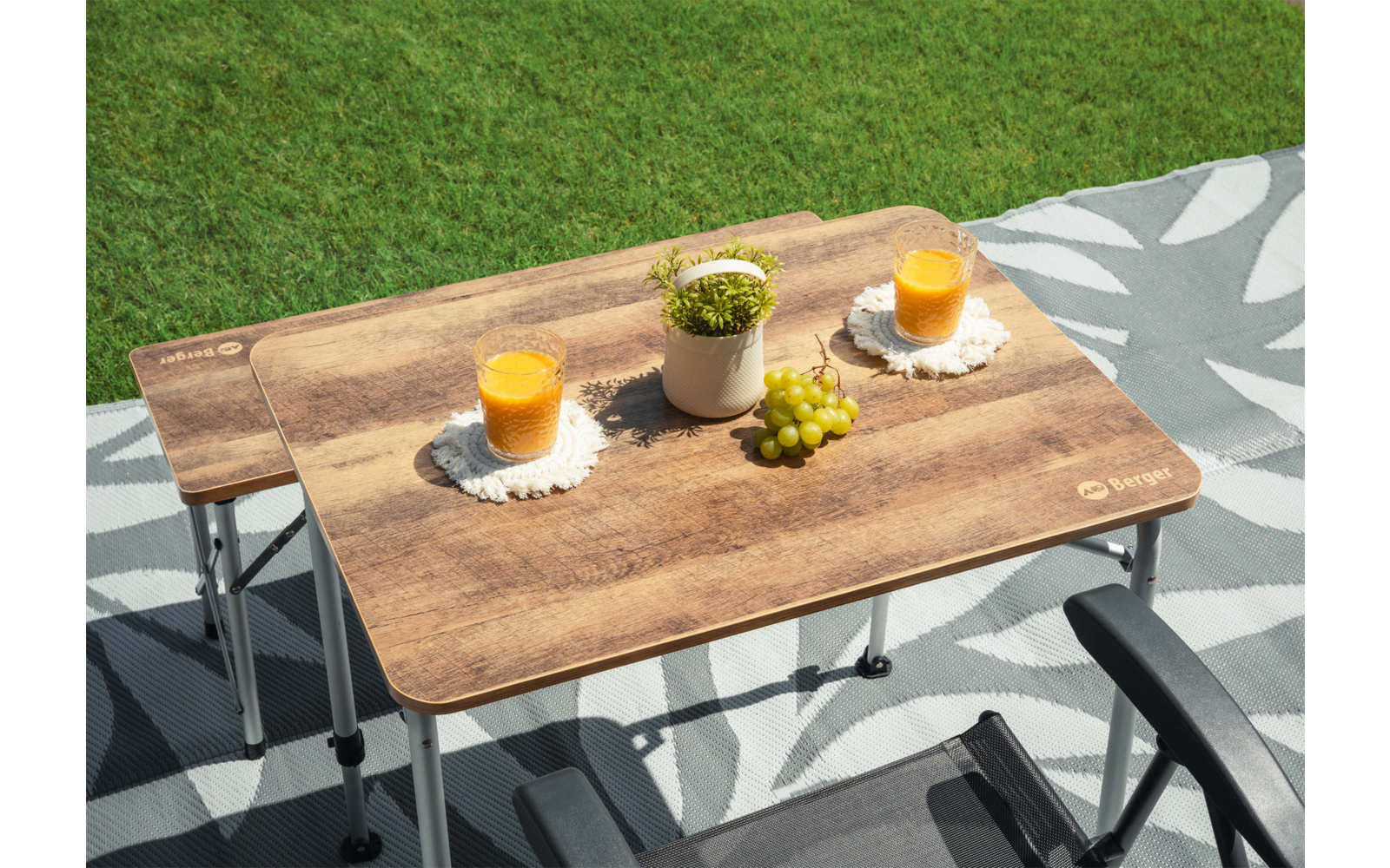 Deux tables pliantes en bois sur un tapis gris à motifs posé sur l'herbe ; dessus, deux verres de jus d'orange sur des sous-verres en crochet, un petit pot blanc avec une plante verte et une grappe de raisins verts ; pieds métalliques pliants et deux accoudoirs de chaises noirs visibles