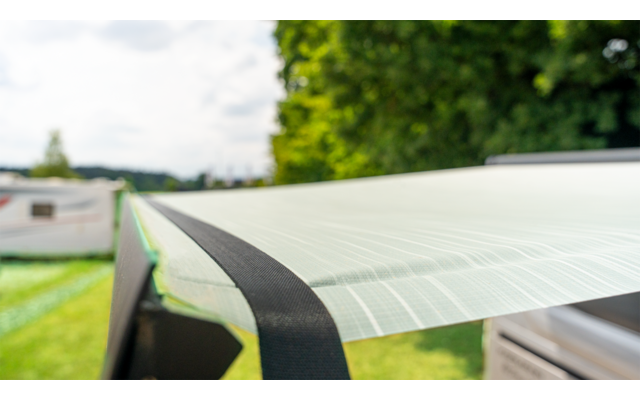 Nahaufnahme eines durchscheinenden, gerippten Kunststoffgewebes, an einem Rahmen mit schwarzem Band befestigt; im Hintergrund unscharfer Wohnwagen und Bäume