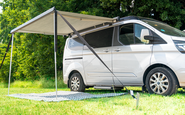 Weißer Campingvan mit ausgefahrener Markise, zwei Stützstangen und Teppich auf Gras vor grünen Bäumen