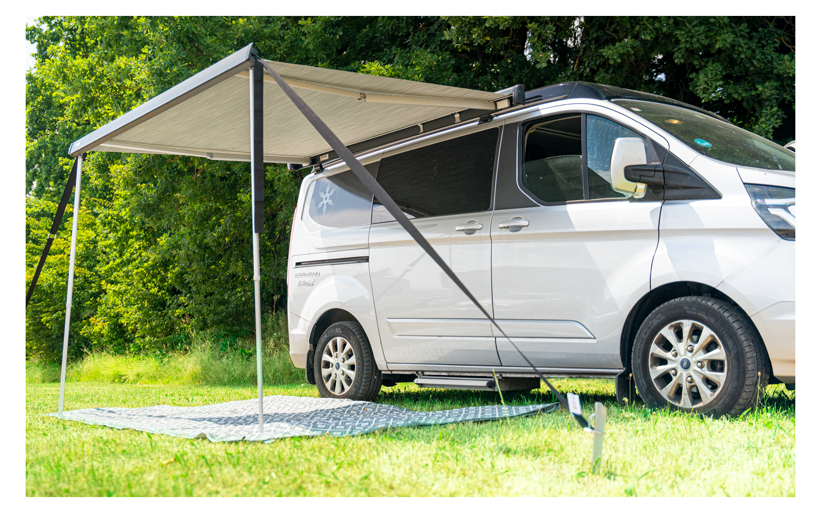 Weißer Campingvan mit ausgefahrener Markise, zwei Stützstangen und Teppich auf Gras vor grünen Bäumen