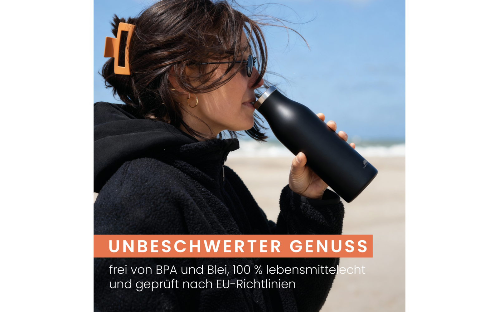 Person trinkt aus einer schwarzen Flasche am Strand, trägt schwarze Jacke, Sonnenbrille, Haarspange und goldenen Creolen; Hintergrund Sand, Meer und blauer Himmel; orangener Banner mit weißem Text 'UNBESCHWERTER GENUSS' und kleinem Text 'frei von BPA