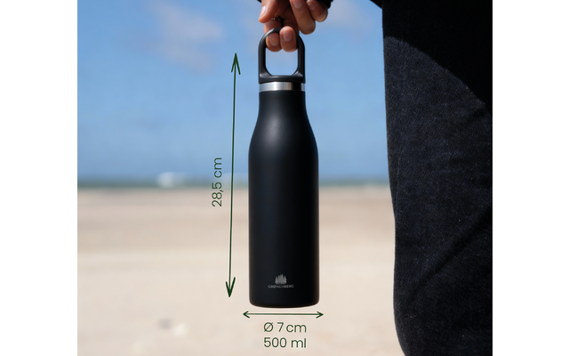 Hand hält schwarze Trinkflasche mit silbernem Verschluss und kleinem Logo, grüne Messpfeile mit Text '28,5 cm' und 'Ø 7 cm 500 ml', Sandstrand, Meer und Himmel im Hintergrund