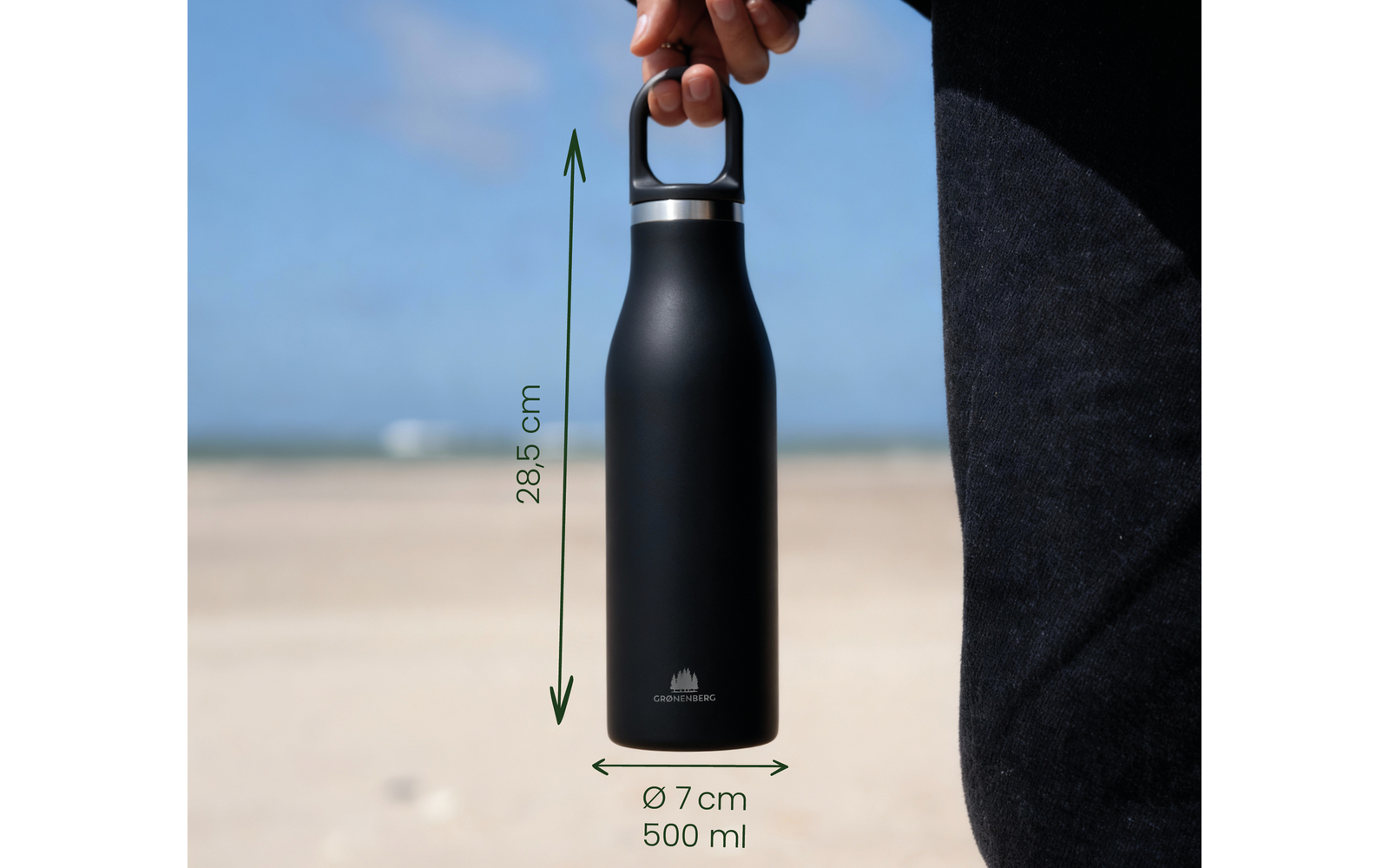 Hand hält schwarze Trinkflasche mit silbernem Verschluss und kleinem Logo, grüne Messpfeile mit Text '28,5 cm' und 'Ø 7 cm 500 ml', Sandstrand, Meer und Himmel im Hintergrund