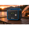 Tragbare Powerstation mit Display, Anschlüssen und Berger-Logo auf einem Holztisch; im Hintergrund ein beleuchtetes Zelt und Berge bei Sonnenuntergang; weiße Pfeile und Beschriftungen auf dem Bild