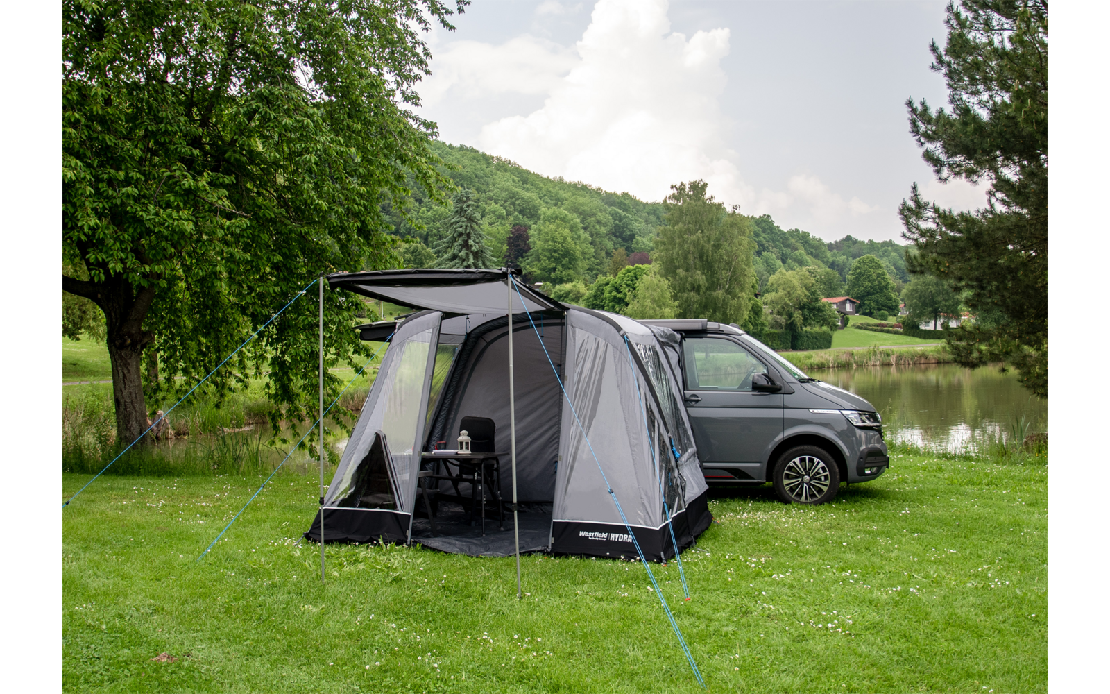 Campingzelt mit Vorzelt neben einem grauen Van auf einer Wiese mit Bäumen im Hintergrund