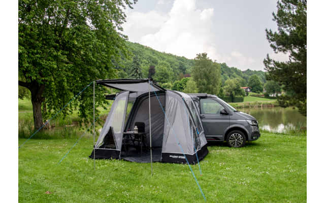 Campingzelt mit Vorzelt neben einem grauen Van auf einer Wiese mit Bäumen im Hintergrund