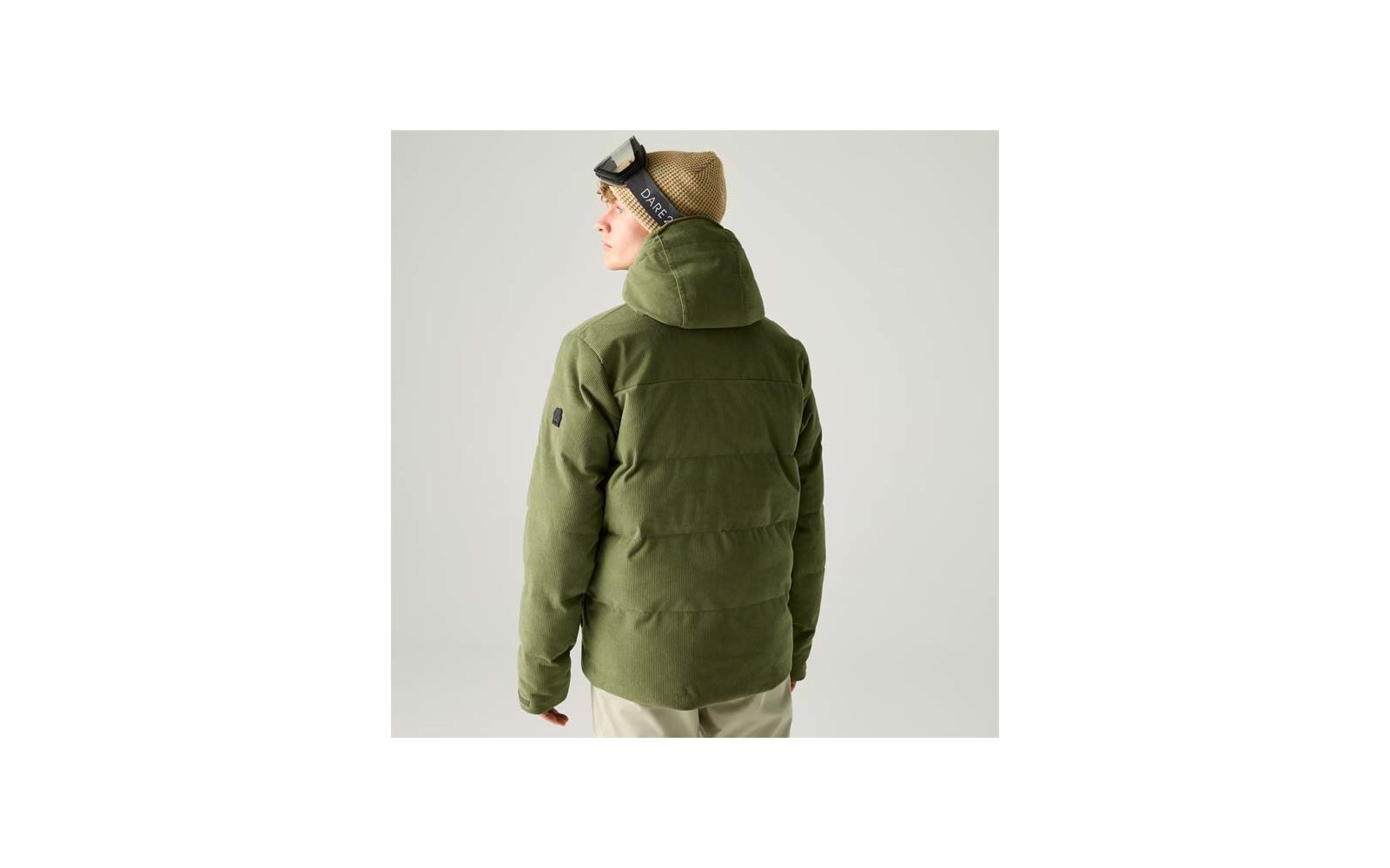 Person von hinten in olivgrüner, gesteppter Jacke mit Kapuze, trägt beige Strickmütze und hochgeschobene Skibrille; hellbeige Hose; heller Hintergrund