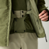 Nahaufnahme des geöffneten Innenbereichs einer olivgrünen Jacke über grünem Pullover und hellen Hosen; elastischer innerer Bund mit Druckknöpfen, Reißverschluss, Pflegeetikett und rechte Hand, die die Jacke hält