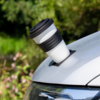 White cup with black lid and black rubber band in a round recess on the hood of a silver car, blurred green background