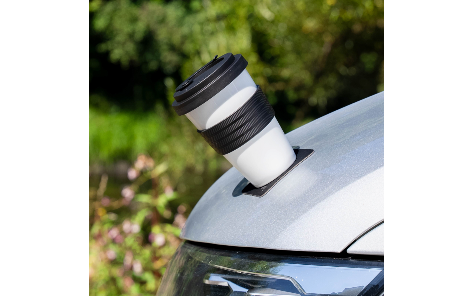 White cup with black lid and black rubber band in a round recess on the hood of a silver car, blurred green background