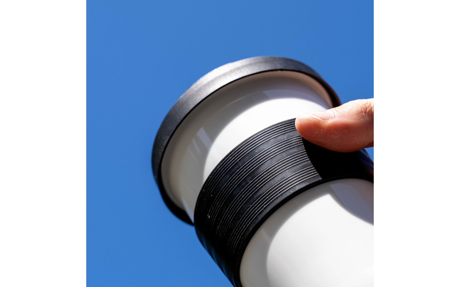 Hand holding a white tumbler with a black ribbed grip against a blue sky