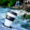 Reusable takeaway coffee cup on a rock at a riverside, background shows a standing wave with a surfer and people on steps