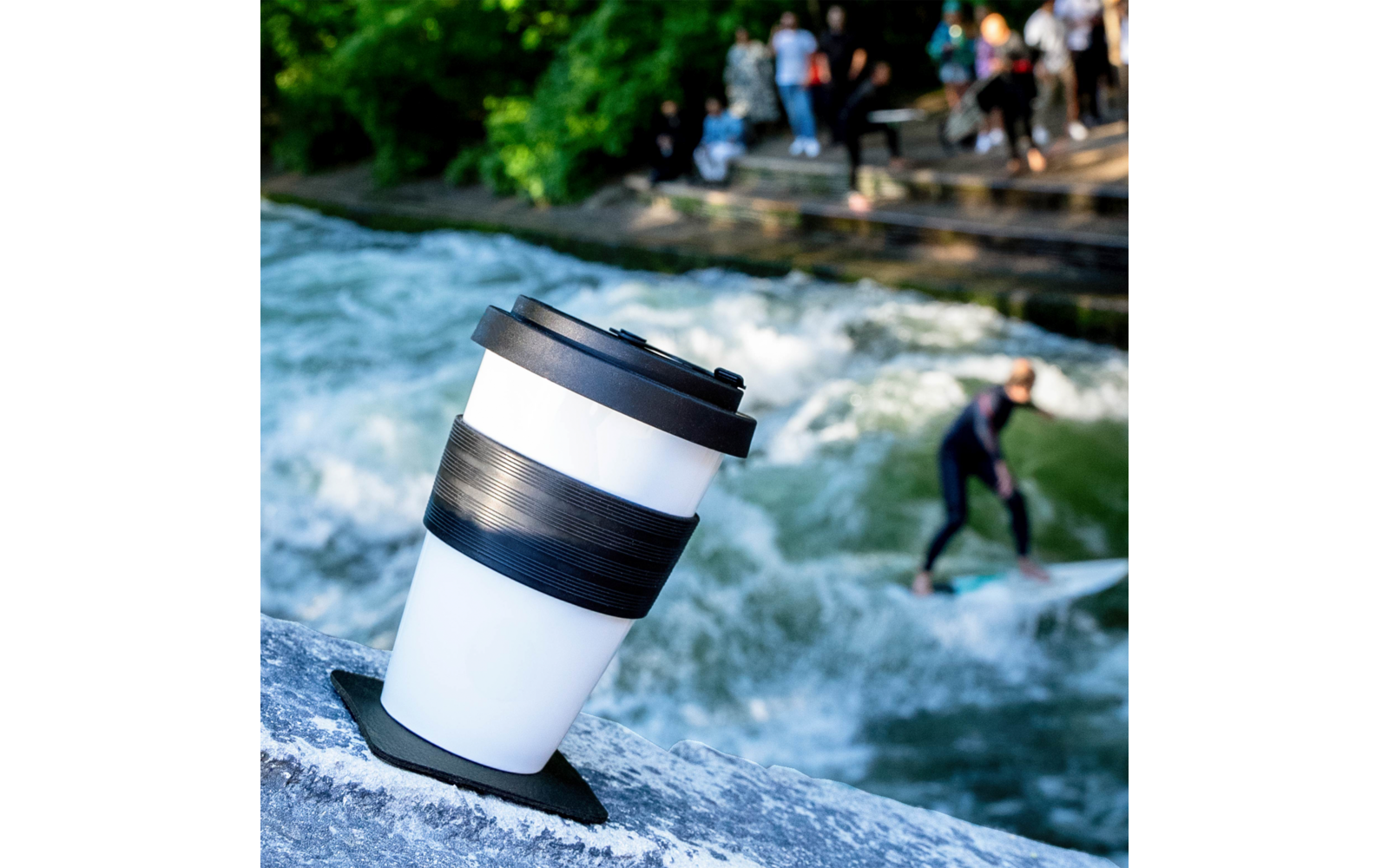 Reusable takeaway coffee cup on a rock at a riverside, background shows a standing wave with a surfer and people on steps