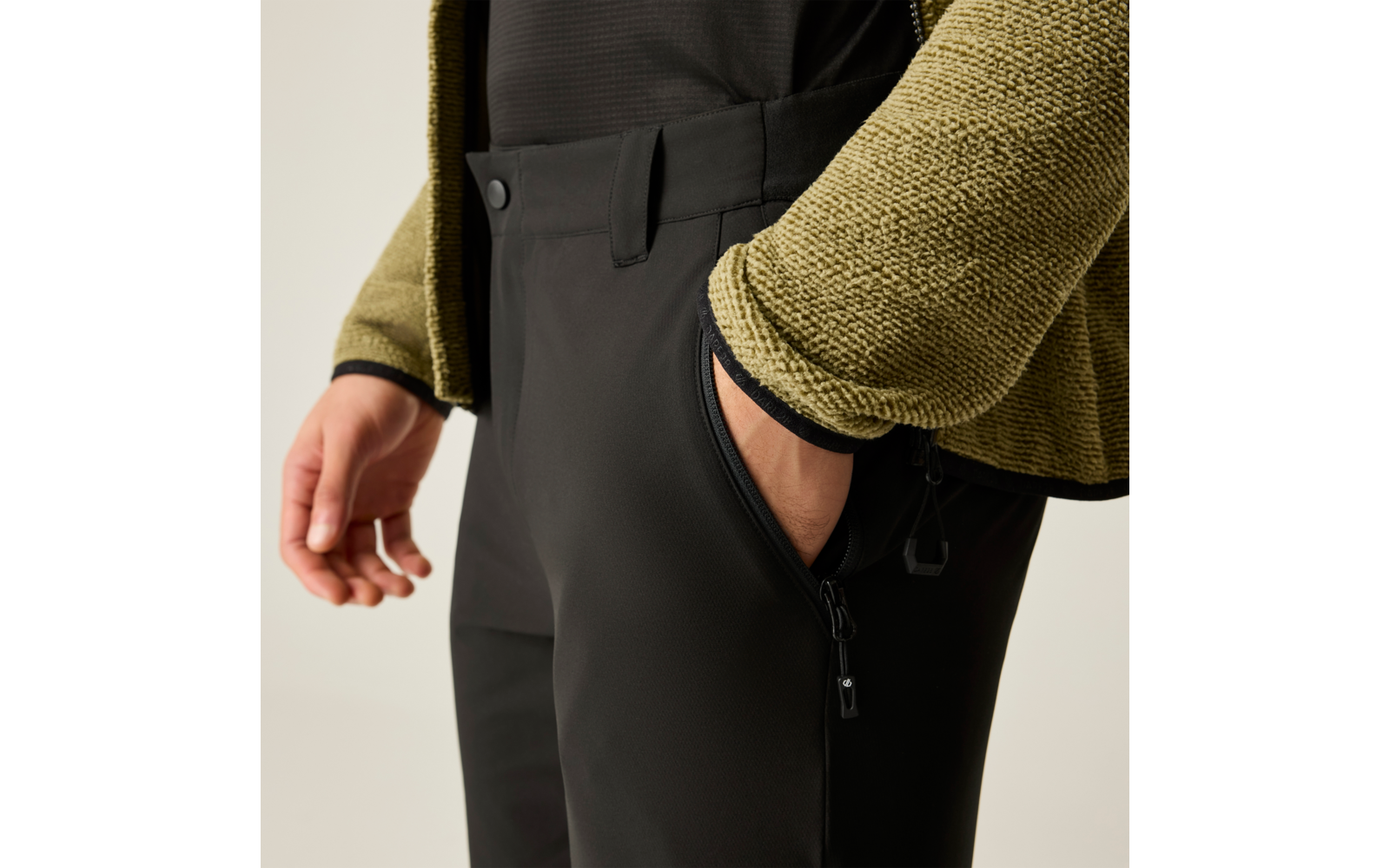 Plano detalle: mano derecha en el bolsillo lateral con cremallera de unos pantalones negros; chaqueta de punto verde y top negro visibles
