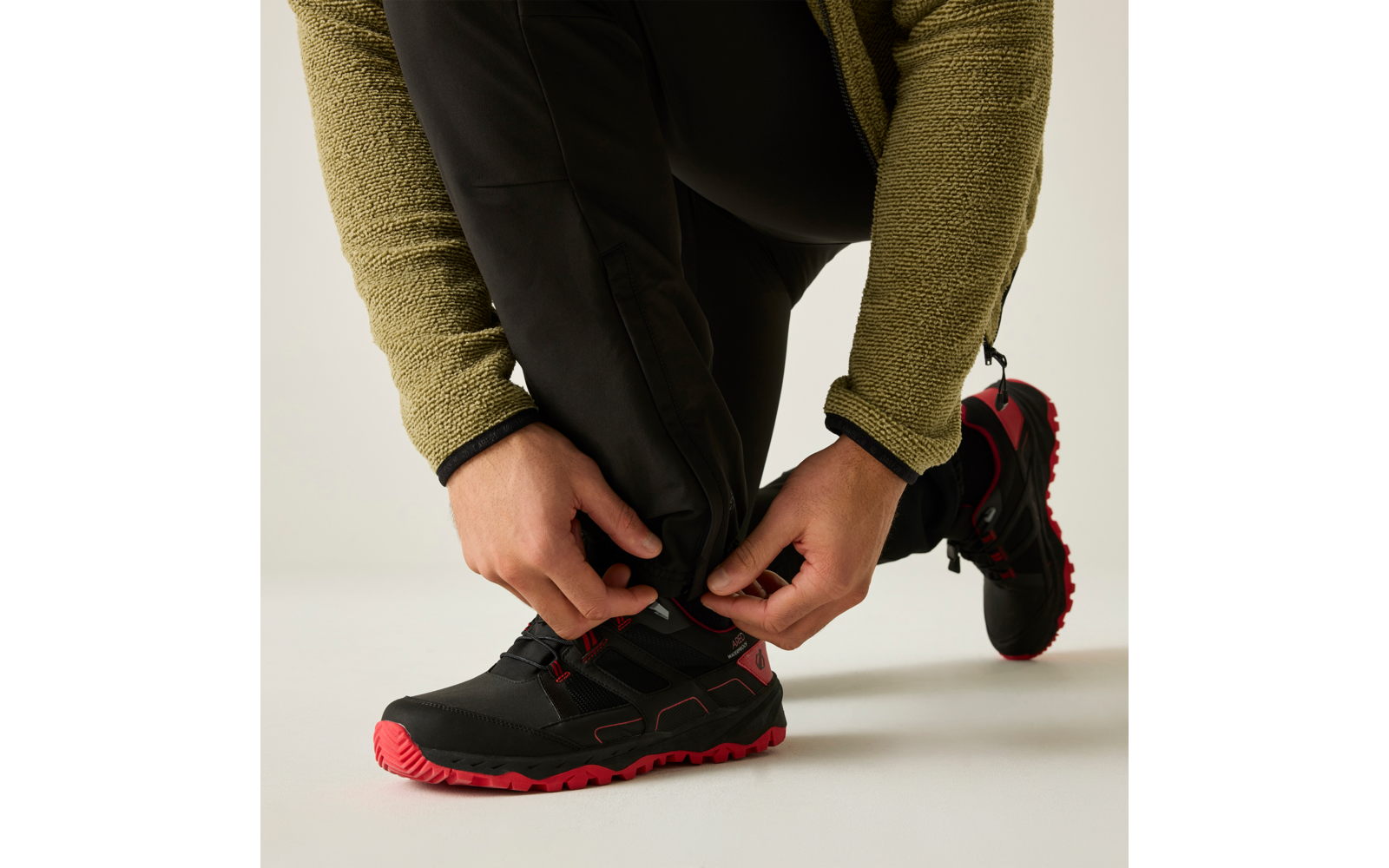 Manos atando los cordones de un zapato negro con suela roja; piernas inferiores con pantalón negro y chaqueta verde visibles