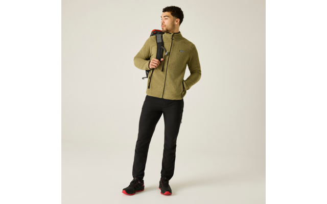 Un hombre de pie con chaqueta de forro polar verde oliva, pantalones negros y zapatillas negras con suelas rojas, lleva una mochila negra sobre el hombro derecho y mira hacia la izquierda