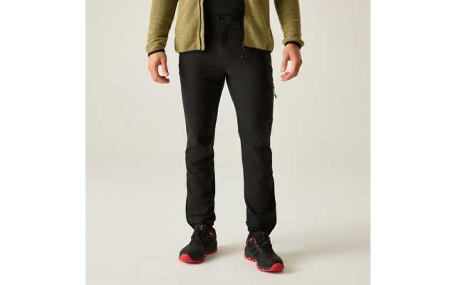 Parte inferior del cuerpo de una persona de pie con chaqueta de forro polar verde oliva, pantalones negros y zapatos negros con suelas rojas sobre fondo claro
