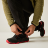 Manos atando los cordones de un zapato negro con suela roja; piernas inferiores con pantalón negro y chaqueta verde visibles