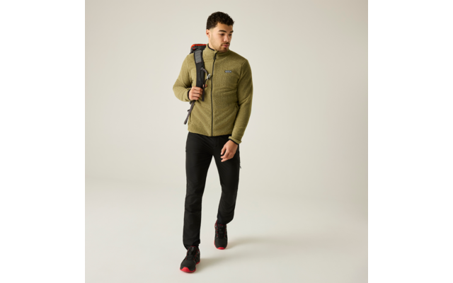 Foto de cuerpo entero de un hombre con pelo corto y barba corta que lleva una chaqueta polar verde oliva, pantalones negros y zapatillas negras con suelas rojas, caminando y con una mochila sobre un hombro sobre un fondo liso claro