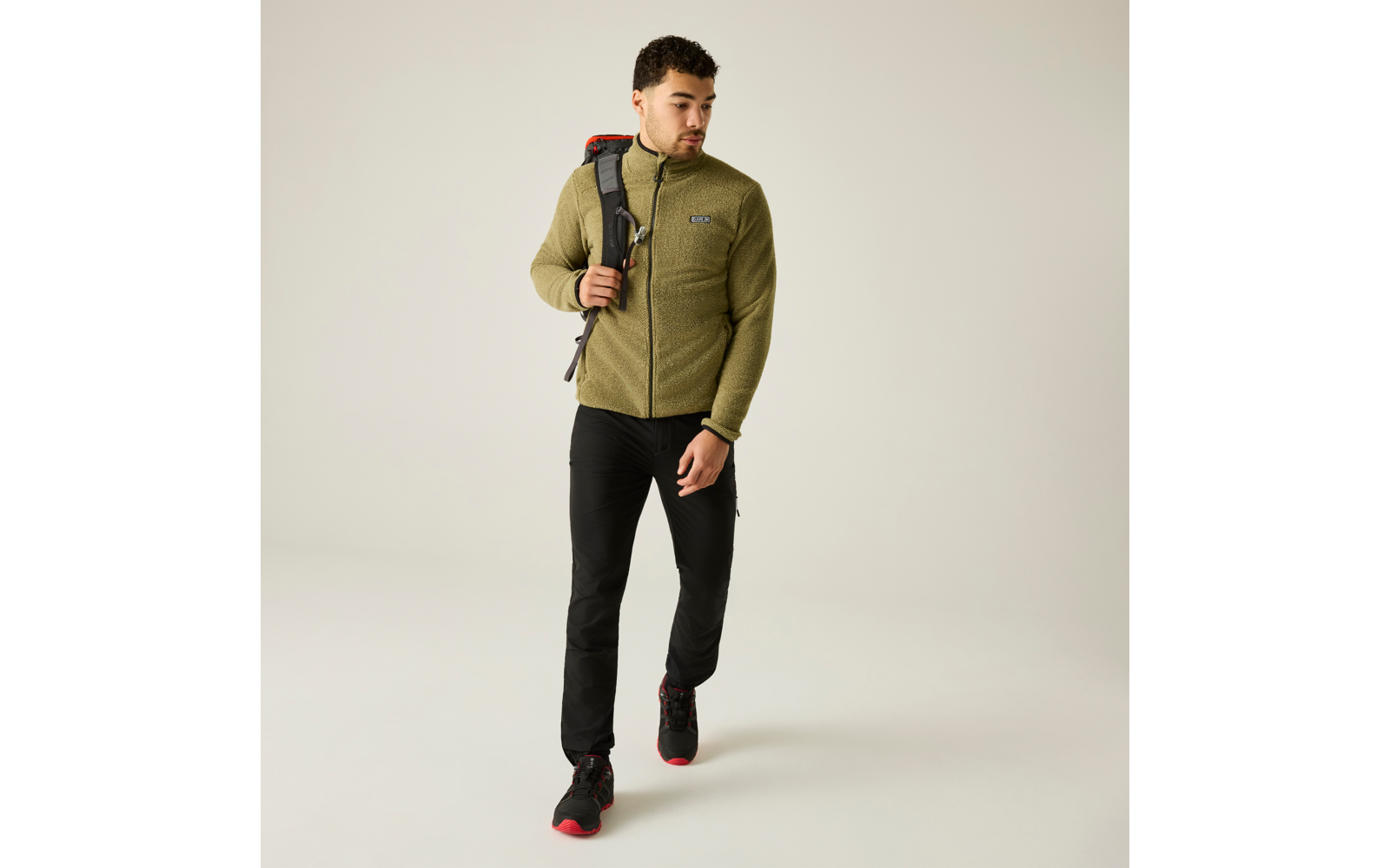 Foto de cuerpo entero de un hombre con pelo corto y barba corta que lleva una chaqueta polar verde oliva, pantalones negros y zapatillas negras con suelas rojas, caminando y con una mochila sobre un hombro sobre un fondo liso claro