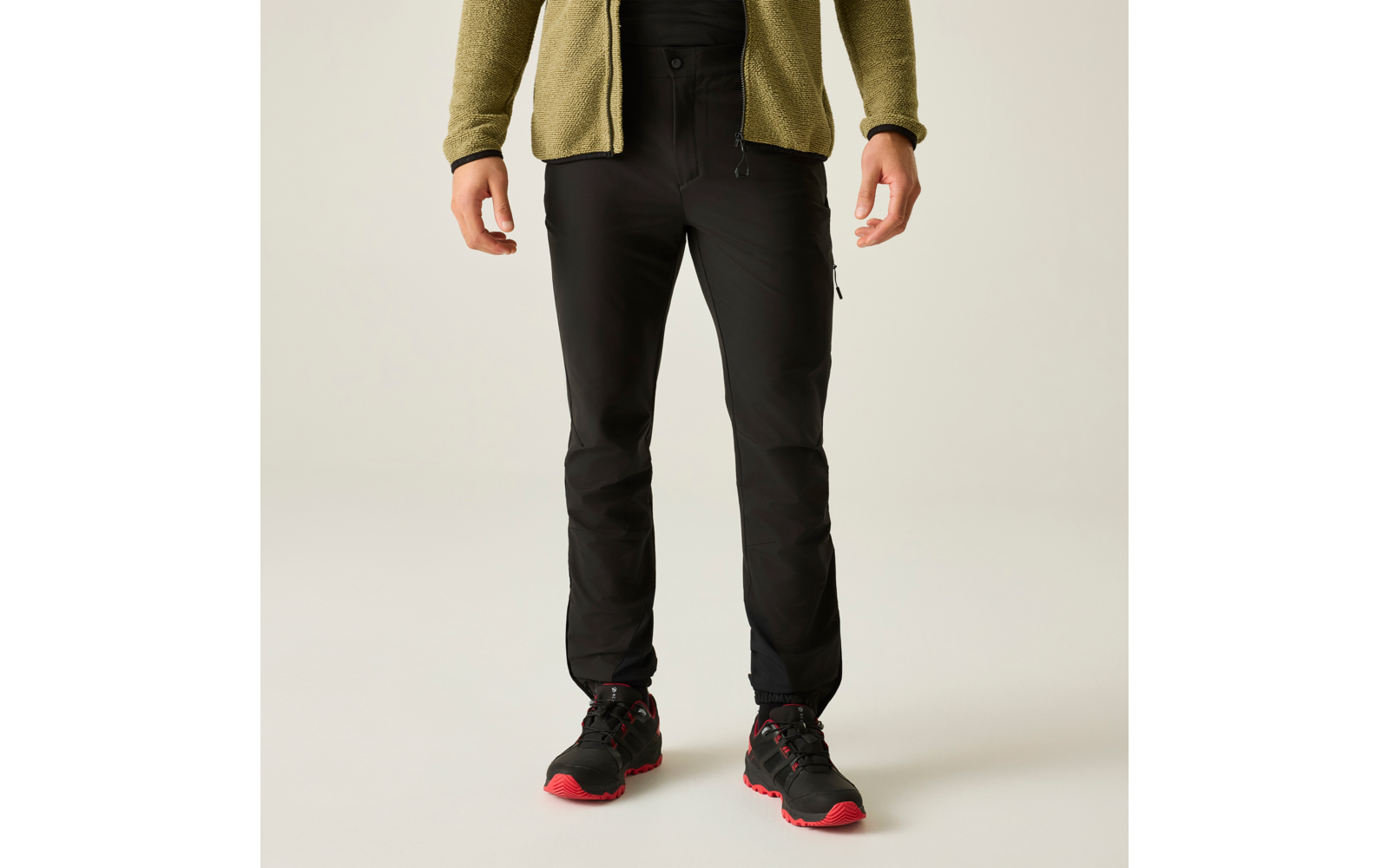 Parte inferior del cuerpo de una persona de pie con chaqueta de forro polar verde oliva, pantalones negros y zapatos negros con suelas rojas sobre fondo claro