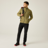 Un hombre de pie con chaqueta de forro polar verde oliva, pantalones negros y zapatillas negras con suelas rojas, lleva una mochila negra sobre el hombro derecho y mira hacia la izquierda