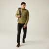 Foto de cuerpo entero de un hombre con pelo corto y barba corta que lleva una chaqueta polar verde oliva, pantalones negros y zapatillas negras con suelas rojas, caminando y con una mochila sobre un hombro sobre un fondo liso claro