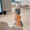 Hand berührt Smartphone auf kleinem Stativ; auf dem Bildschirm ist das Gesicht einer Person im Videoanruf; Schreibtisch mit Monitor und Zeitschriften im Hintergrund