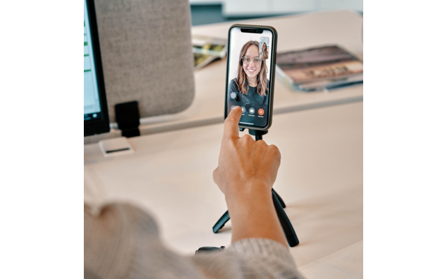 Hand berührt Smartphone auf kleinem Stativ; auf dem Bildschirm ist das Gesicht einer Person im Videoanruf; Schreibtisch mit Monitor und Zeitschriften im Hintergrund