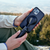 Zwei Hände halten ein Smartphone in schwarzer Hülle, befestigt an einem schwarzen Halter mit rundem Gelenk; verschwommene Waldlandschaft im Hintergrund