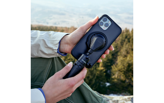 Zwei Hände halten ein Smartphone in schwarzer Hülle, befestigt an einem schwarzen Halter mit rundem Gelenk; verschwommene Waldlandschaft im Hintergrund